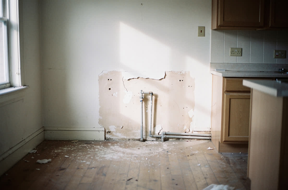 Stripped fixer-upper kitchen with cabinets removed from one wall, bare plaster and capped pipework