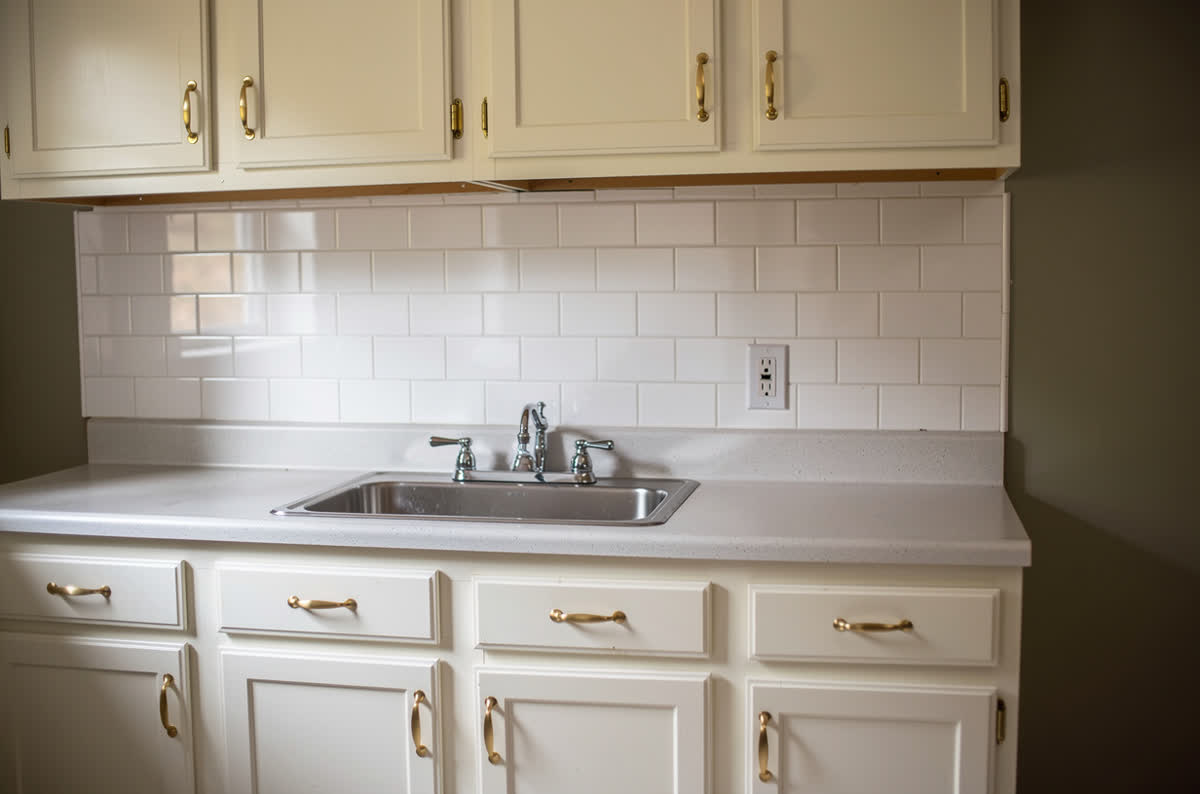 The same kitchen after an 8,000 dollar refresh: cabinets repainted warm white, brass handles, subway tile and a new sink