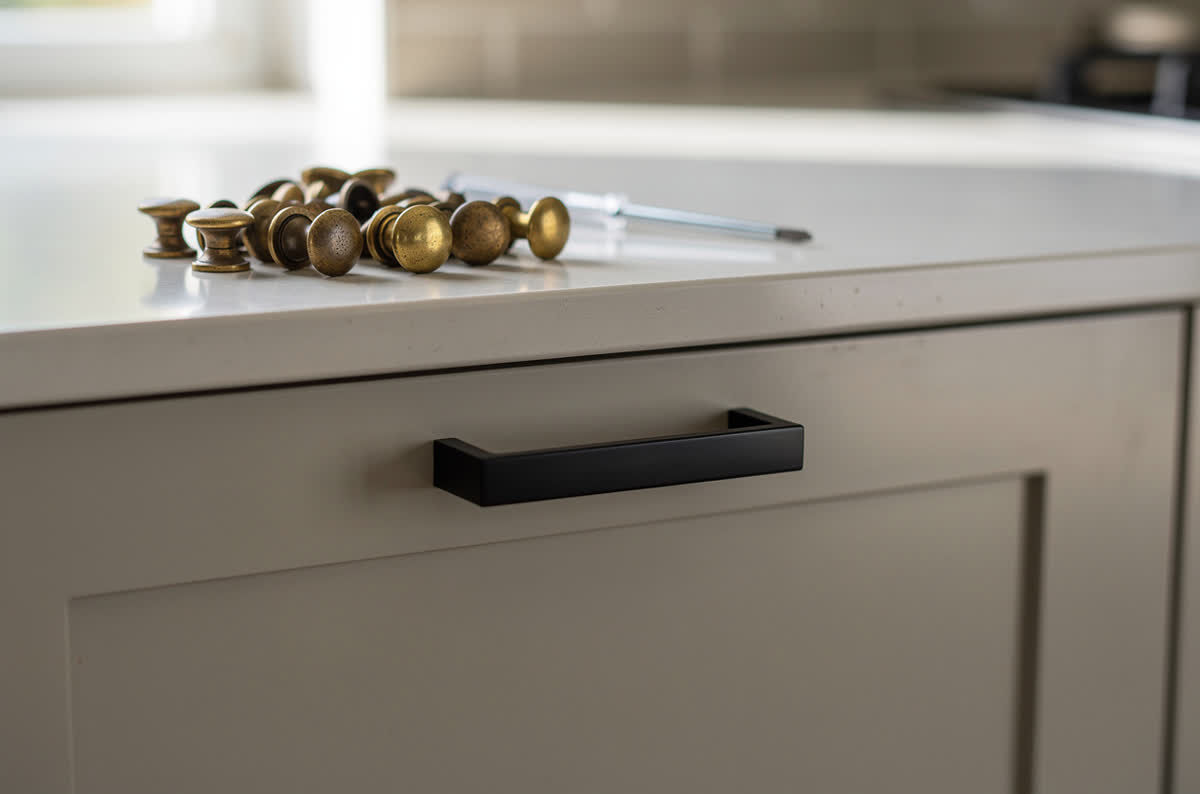 New matte black bar pull on a drawer front beside a pile of old brass knobs and a screwdriver