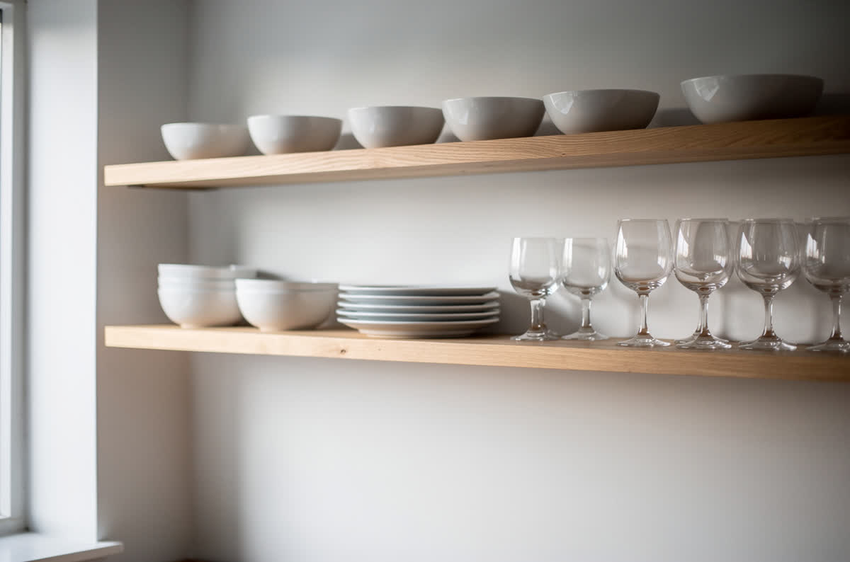 Kitchen wall with no upper doors, three open wooden shelves holding crockery and glassware